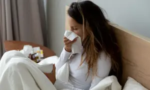 Kobieta w łóżku z chusteczką przy nosie i kubkiem w dłoni.