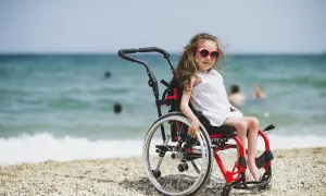 Dziewczynka w okularach przeciwsłonecznych siedzi na plaży w wózku inwalidzkim. W tle widać morze i ludzi kąpiących się.