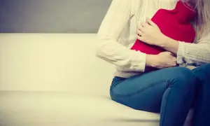 Kobieta siedzi na kanapie, przytulając czerwoną termofor. Czuje ból brzucha po jedzeniu i szuka ulgi.