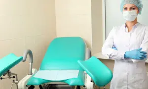 Lekarka w maseczce i rękawiczkach stoi obok fotela ginekologicznego. Wszystko gotowe na wizytę na cito.