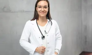 Lekarka w białym fartuchu ze stetoskopem na szyi, na tle szarej ściany.