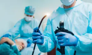 Lekarz w niebieskich rękawiczkach trzyma endoskop. W tle pacjent i druga lekarka. Procedura gastroskopii jest szybka.
