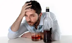 Mężczyzna z ręką na czole obok butelki i szklanki z alkohoholem.