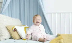 Uśmiechnięte dziecko w różowym pajacyku siedzi na łóżku, otoczone poduszkami i niebieskim baldachimem. To radosny moment, kiedy dziecko siada samodzielnie.