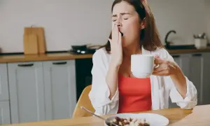 Kobieta z kubkiem kawy ziewa, czując senność po jedzeniu. Na stole miska płatków.