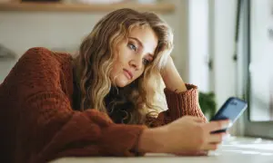 Młoda kobieta w brązowym swetrze, z ręką opartą o głowę, patrzy na telefon. Jej wyraz twarzy sugeruje anhedonię, brak radości z życia.