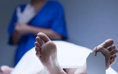 Stopy z metką identyfikacyjną, widoczne plamy opadowe. W tle osoba w niebieskim fartuchu zakłada rękawiczki.