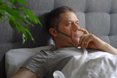 Mężczyzna w masce tlenowej leży w łóżku, podłączony do aparatu do inhalacji.