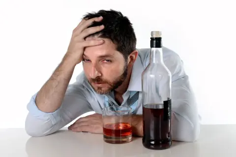 Mężczyzna z ręką na czole obok butelki i szklanki z alkohoholem.