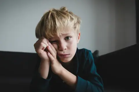 Chłopiec z blond włosami opiera głowę na dłoniach, patrząc w obiektyw z zamyślonym wyrazem twarzy.