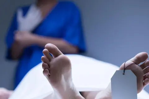 Stopy z metką identyfikacyjną, widoczne plamy opadowe. W tle osoba w niebieskim fartuchu zakłada rękawiczki.