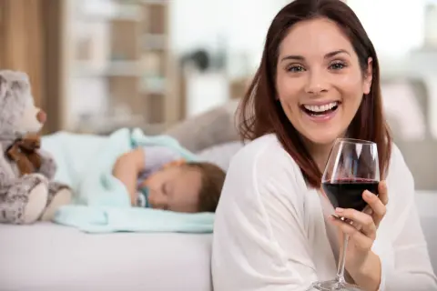 Uśmiechnięta kobieta z kieliszkiem czerwonego wina obok śpiącego dziecka. Czy można pić alkohol przy kamicy żółciowej?