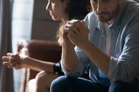Para siedzi obok siebie, mężczyzna z rękami złożonymi w geście modlitwy, kobieta odwrócona.