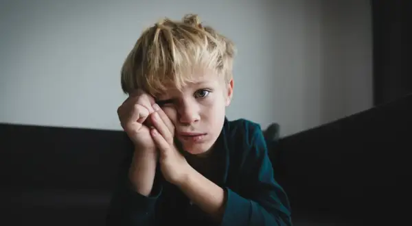 Chłopiec z blond włosami opiera głowę na dłoniach, patrząc w obiektyw z zamyślonym wyrazem twarzy.