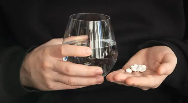 Dwie dłonie trzymają szklankę wody i garść białych tabletek. Zastanawiasz się, czy można pić alkohol po Ibupromie?