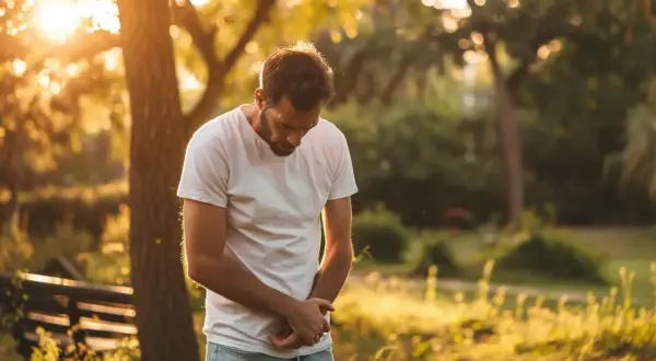 Mężczyzna w białej koszulce trzyma się za brzuch, zgięty w pół. Słońce przebija się przez drzewa, tworząc ciepłą, złotą poświatę. Może to być symbol bólu związanego z chorobą Leśniowskiego-Crohna.