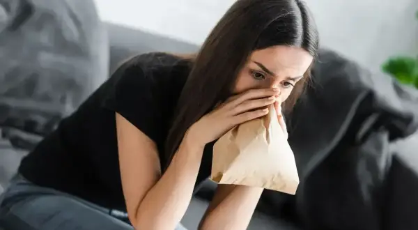 Kobieta siedzi na kanapie, trzymając papierową torbę przy ustach i nosie, próbując opanować **hiperwentylację**.