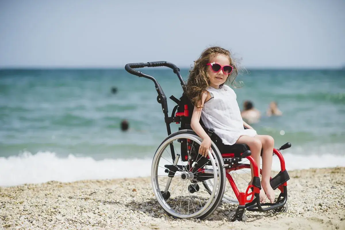 Dziewczynka w okularach przeciwsłonecznych siedzi na plaży w wózku inwalidzkim. W tle widać morze i ludzi kąpiących się.