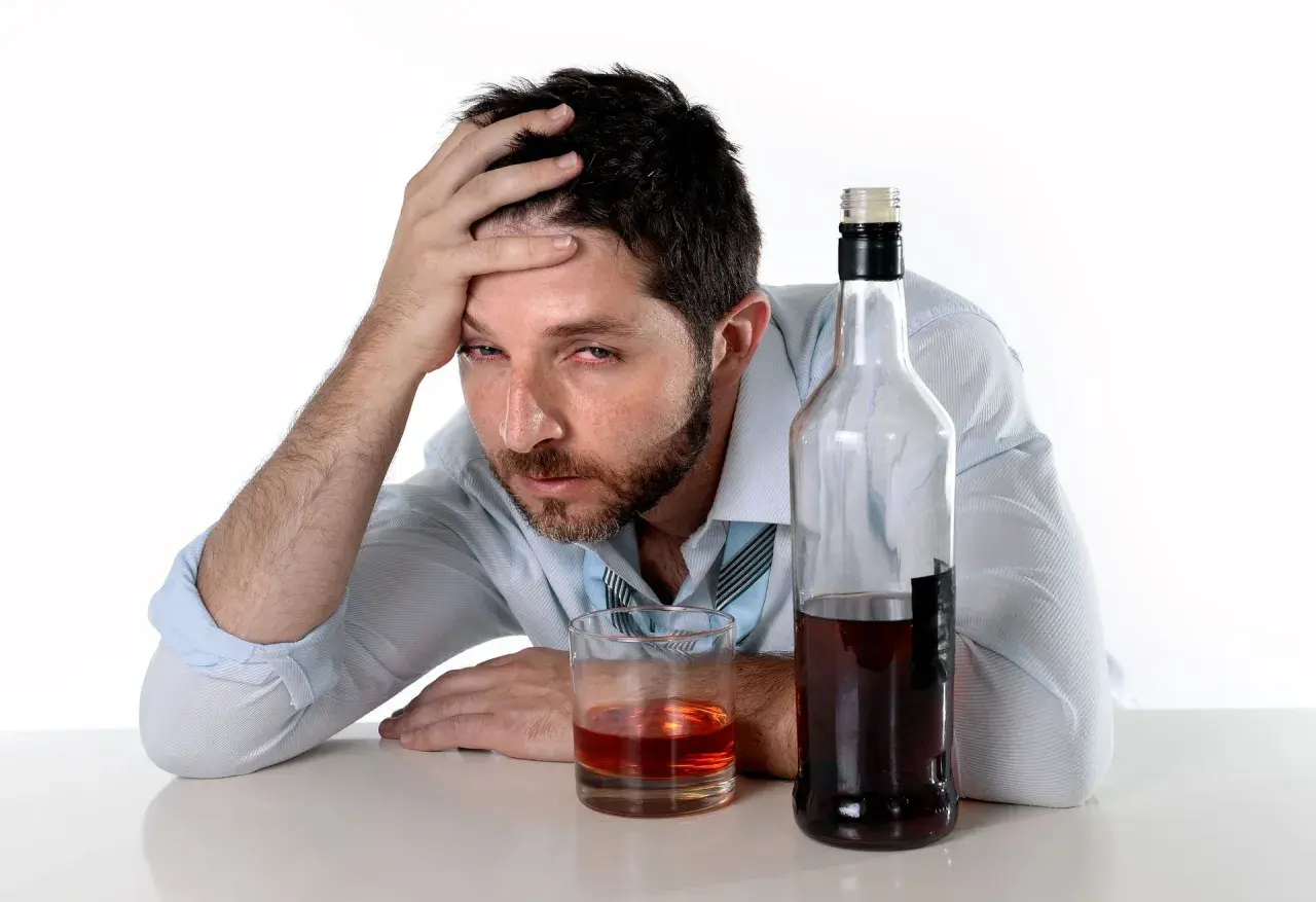 Mężczyzna z ręką na czole obok butelki i szklanki z alkohoholem.