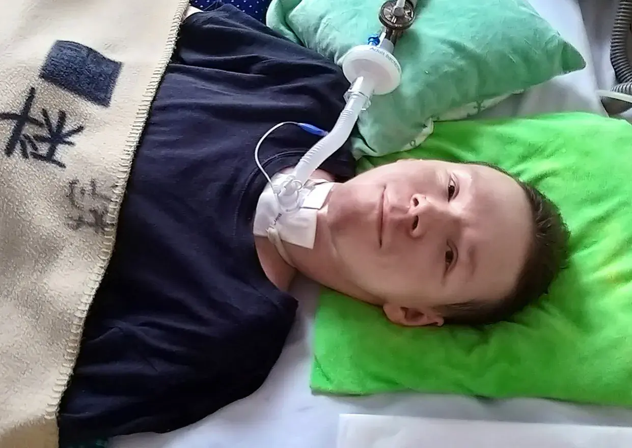 Chłopiec leży w łóżku z rurką tracheostomijną i podłączonym respiratorem.
