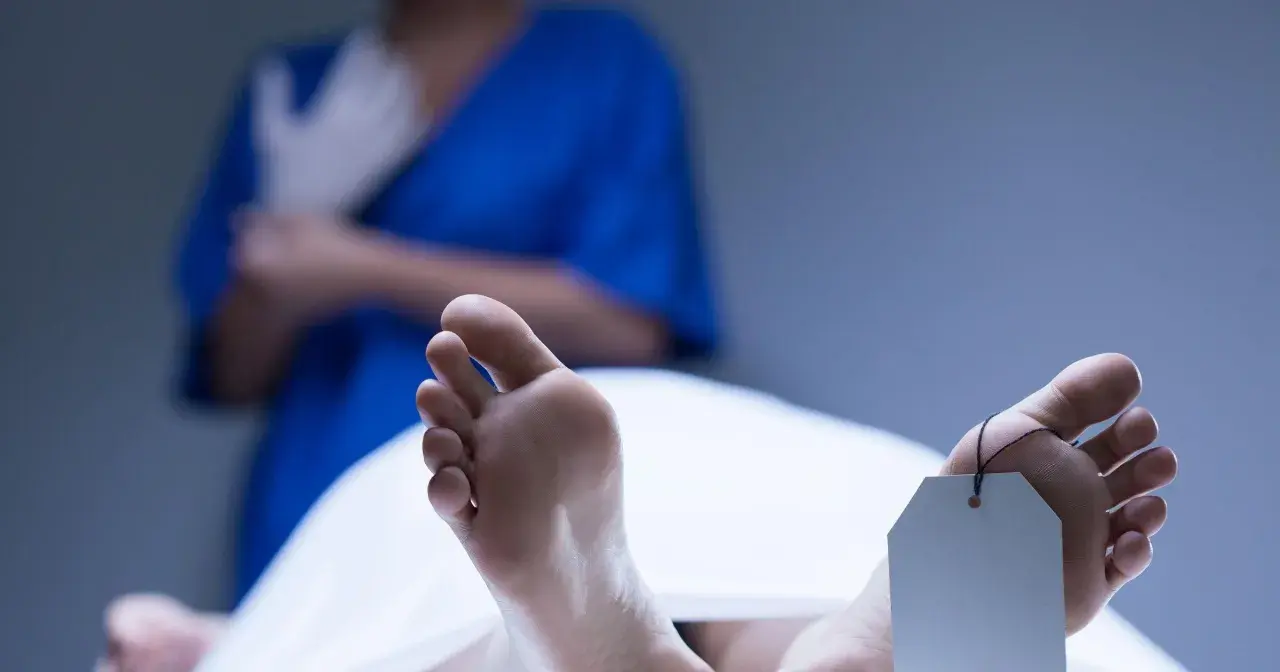 Stopy z metką identyfikacyjną, widoczne plamy opadowe. W tle osoba w niebieskim fartuchu zakłada rękawiczki.