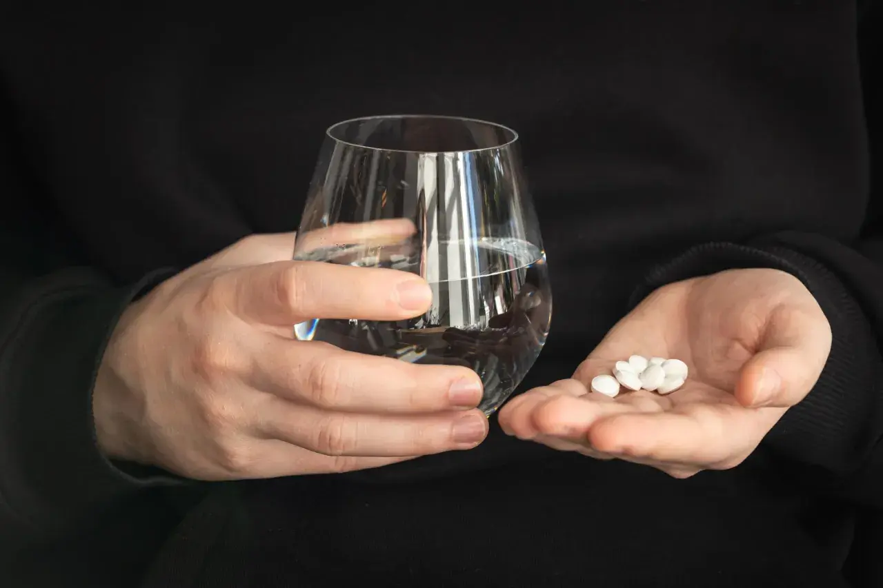 Dwie dłonie trzymają szklankę wody i garść białych tabletek. Zastanawiasz się, czy można pić alkohol po Ibupromie?
