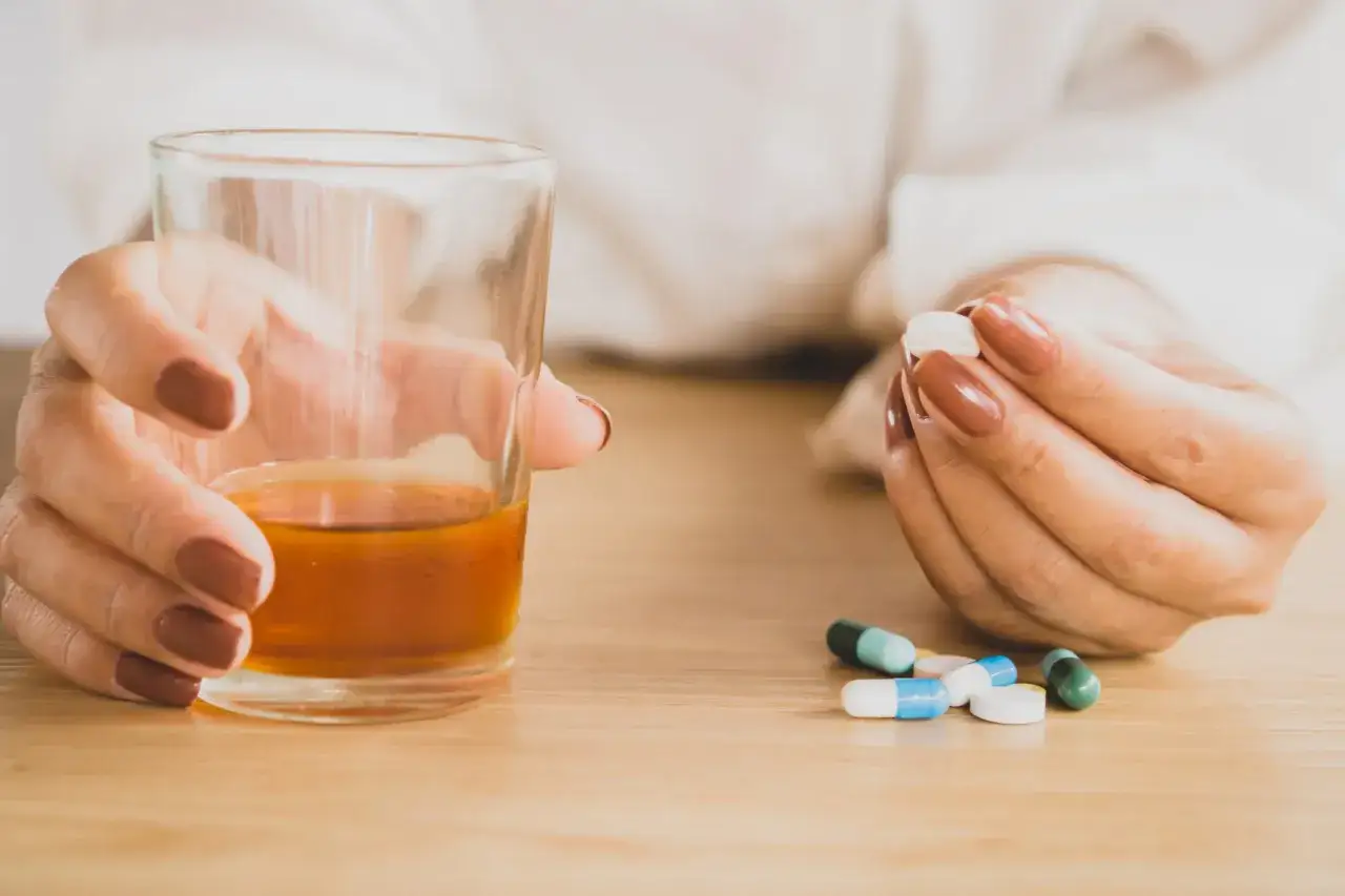 Kobieta trzyma szklankę z bursztynowym płynem i tabletkę, obok leżą kapsułki. Czy nebilet 5 mg można pić alkohol?