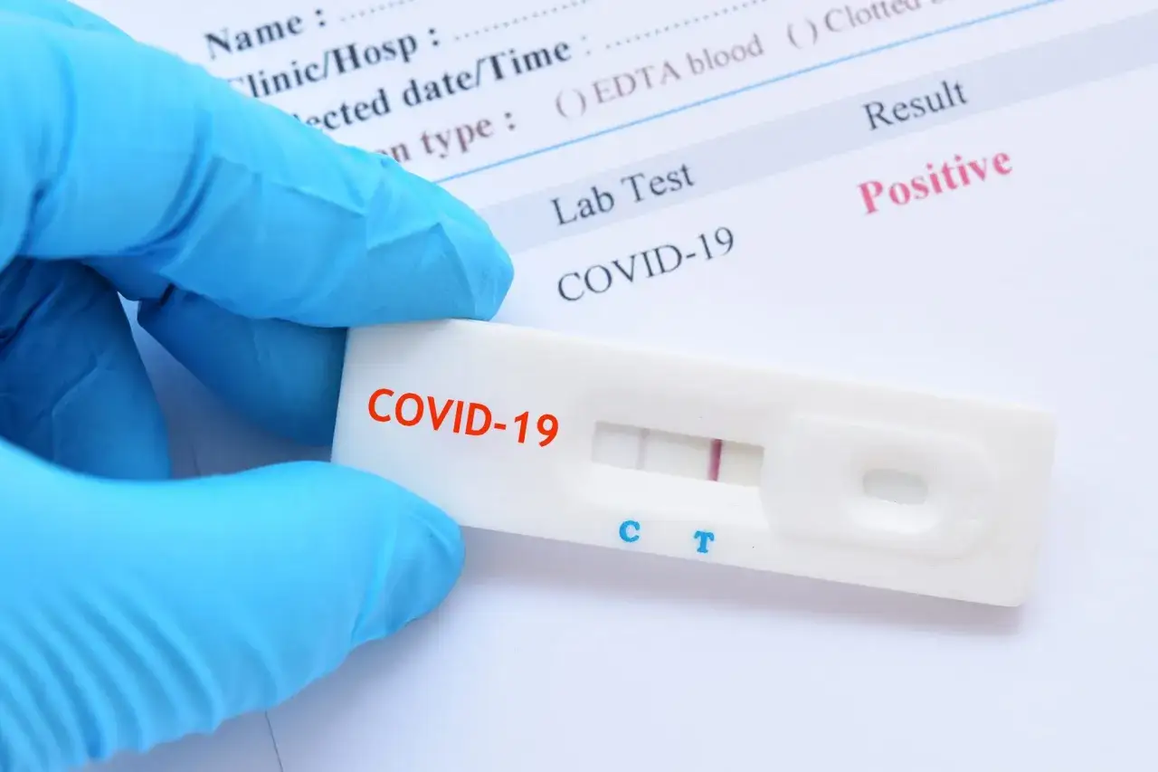 Ręka w niebieskiej rękawiczce trzyma test antygenowy COVID-19 z dwoma widocznymi paskami, co oznacza pozytywny wynik.