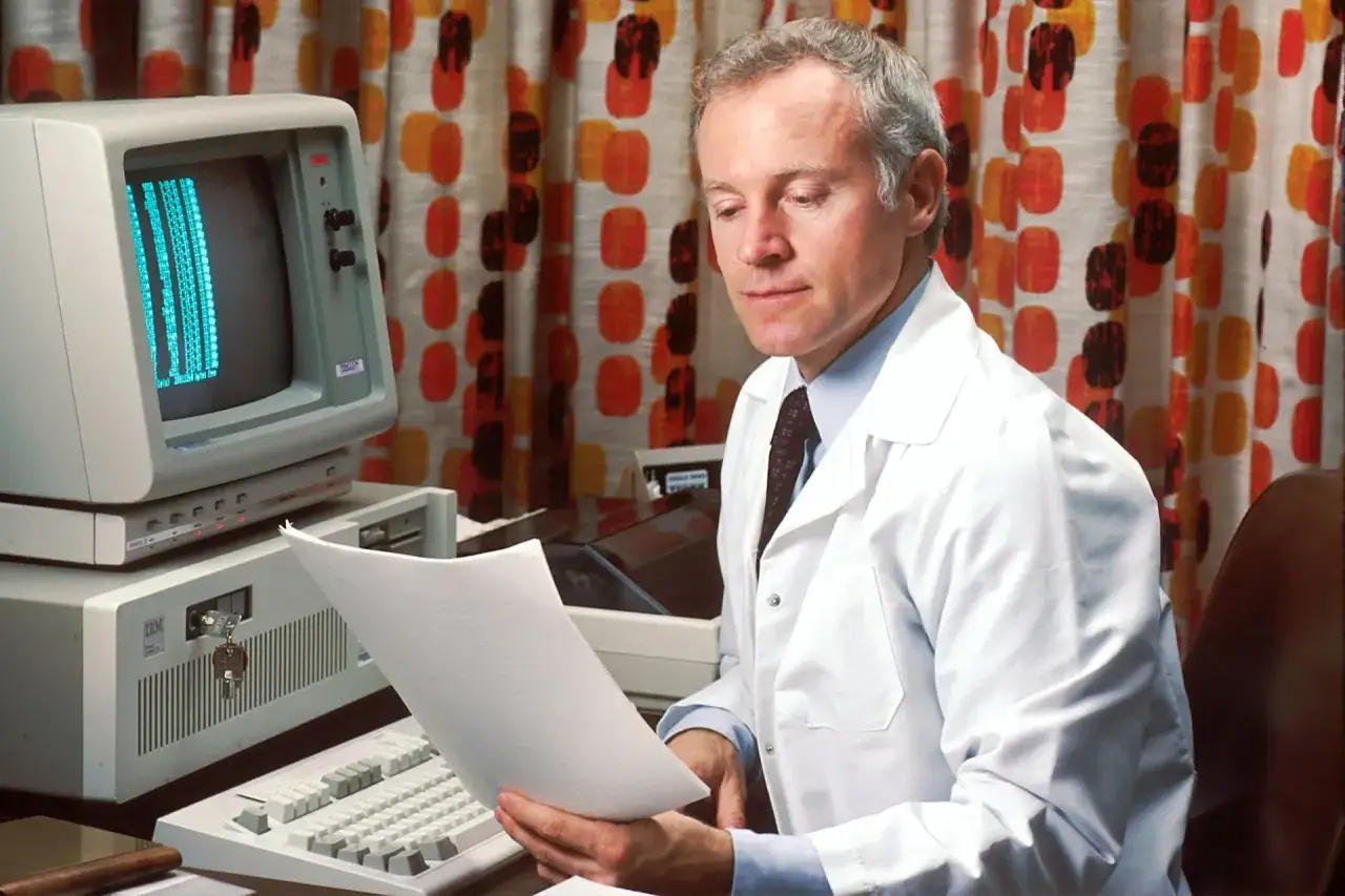Mężczyzna w białym kitlu pochyla się nad dokumentem, obok starego komputera IBM.
