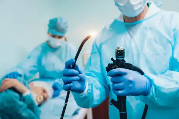 Lekarz w niebieskich rękawiczkach trzyma endoskop. W tle pacjent i druga lekarka. Procedura gastroskopii jest szybka.