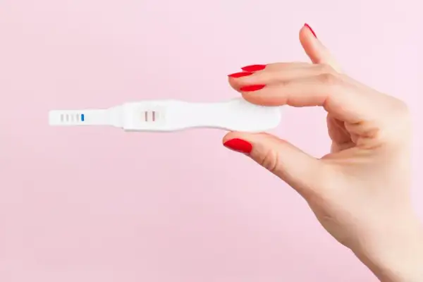 Dłoń z czerwonymi paznokciami trzyma biały test ciążowy z dwoma różowymi paskami.