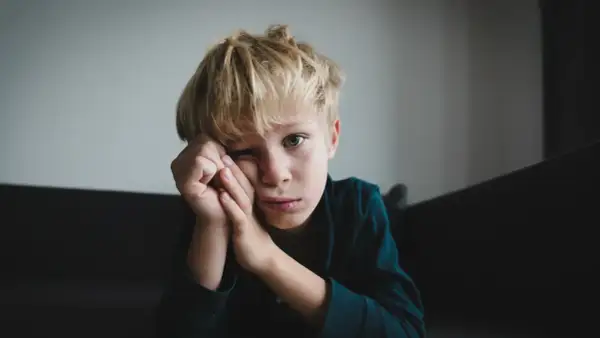 Chłopiec z blond włosami opiera głowę na dłoniach, patrząc w obiektyw z zamyślonym wyrazem twarzy.